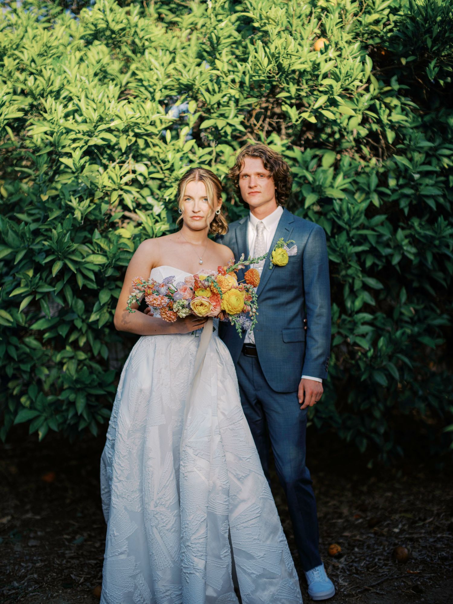 A wedding couple poses in front of lush green foliage wearing a white gown and navy suit with colorful bouquet.
