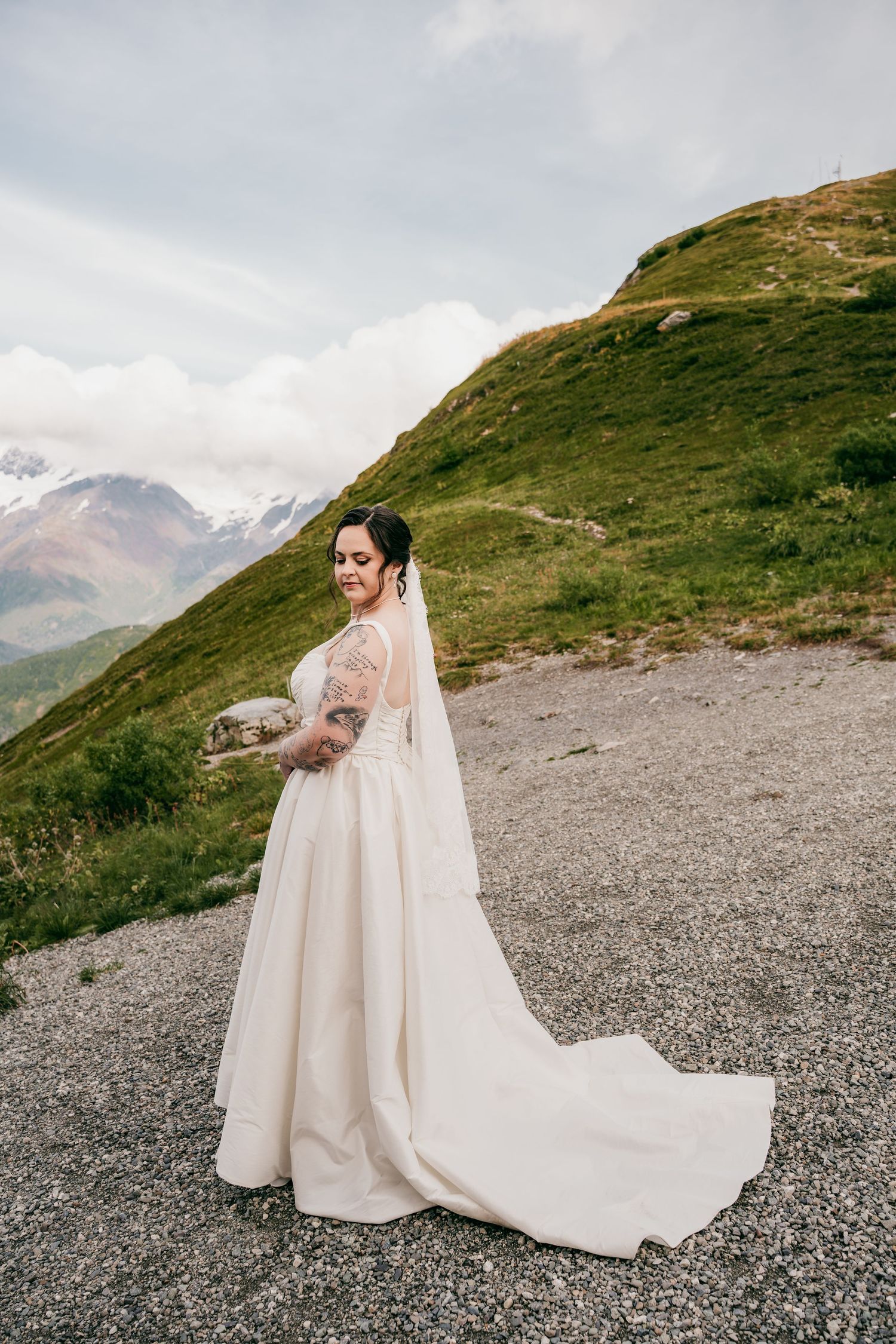 Bride in white lace gown with veil poses on mountain path with green hills and snowy peaks in background.