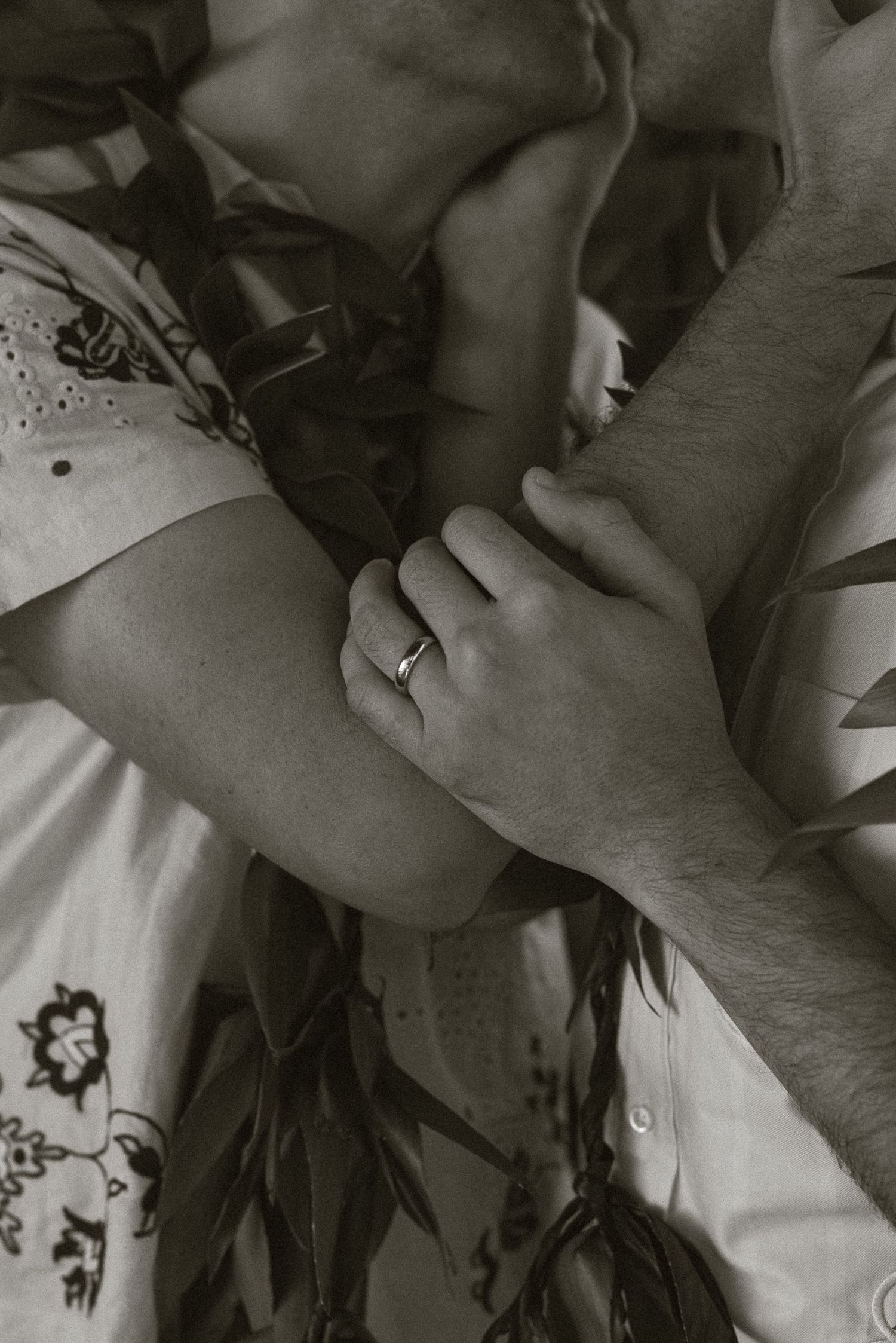 Close-up of two grooms holding hands with a wedding ring visible, surrounded by tropical lei flowers.