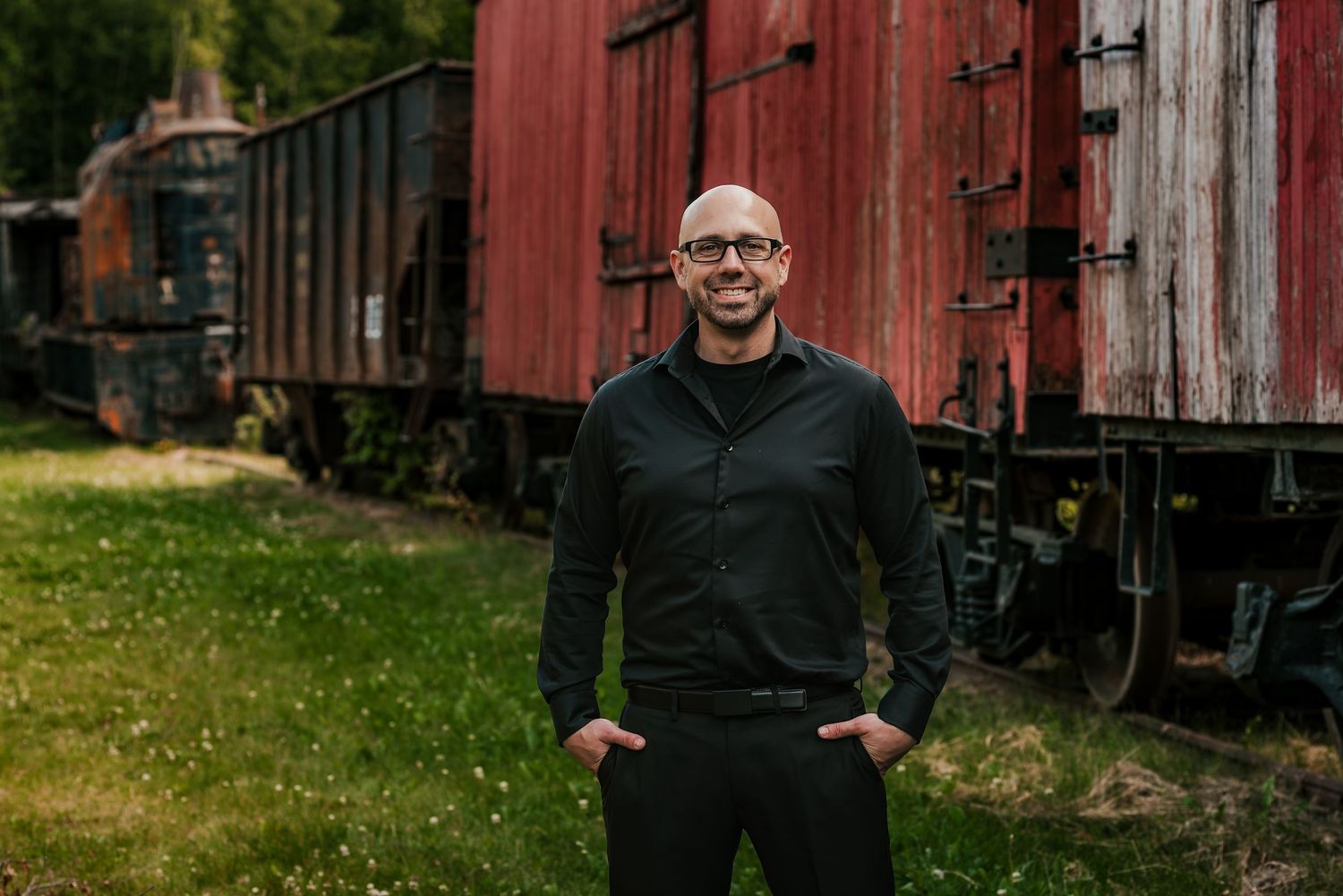 Bald man in black outfit smiling outdoors near vintage red train cars on green grass in wasilla alaska