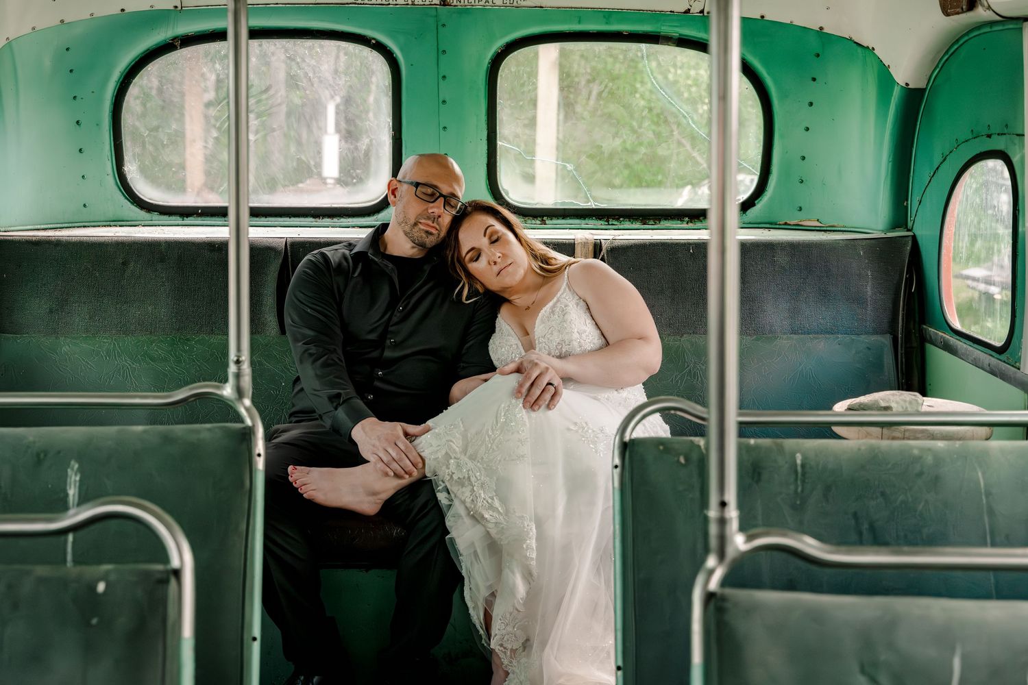 Bride and groom sitting intimately on vintage green bus seats, bride in white gown, groom in black attire.