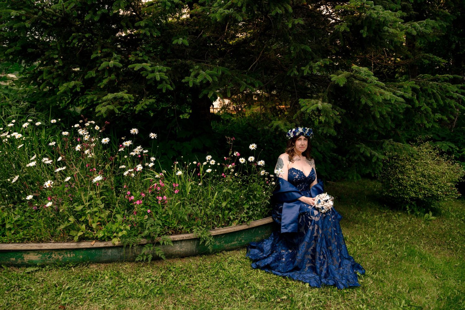 Woman in blue Victorian gown with floral crown poses in lush garden near colorful flowers and evergreen trees.