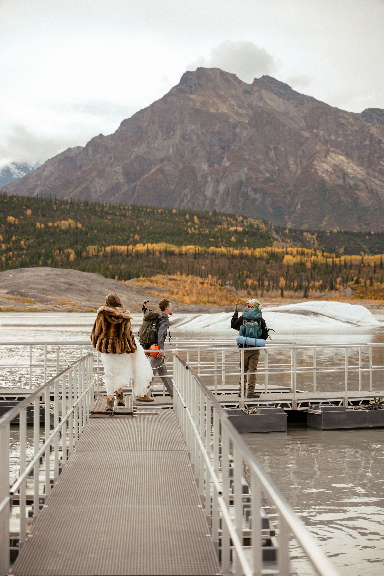 Group of hikers with backpacks walking on a dock toward a glacier with autumn mountains in the background.