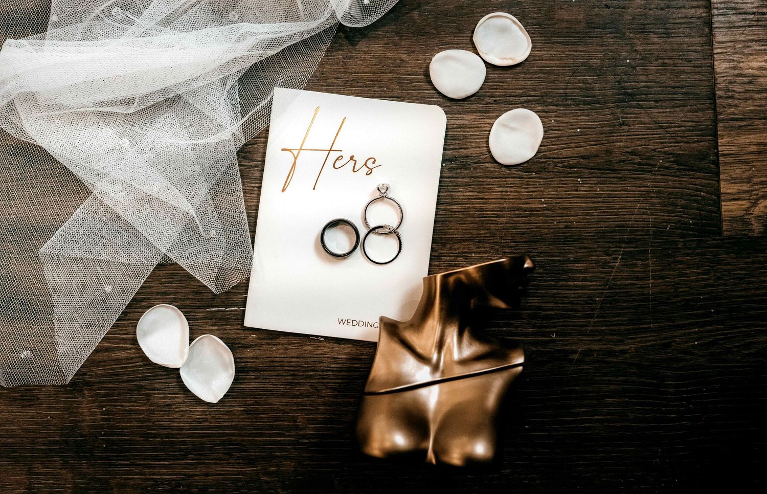Wedding vows card with gold rings, white veil, and tea lights arranged on dark wooden surface.