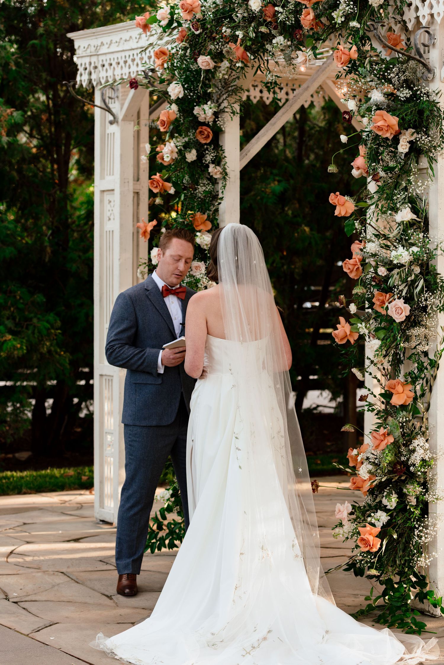 Bride and groom exchange vows under a floral arch adorned with peach roses and greenery at an outdoor wedding ceremony.