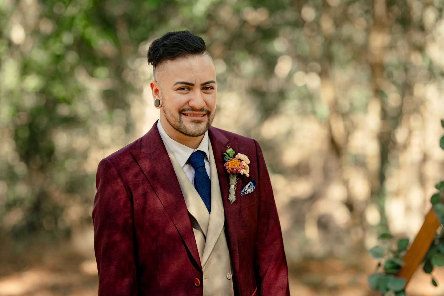 Smiling groom in burgundy plaid suit with floral boutonniere and navy tie, standing outdoors among trees.