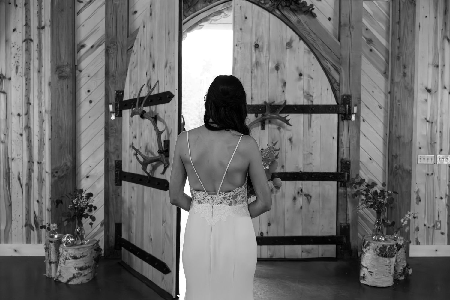 Bride in elegant backless wedding gown standing before rustic wooden barn doors in black and white photo.