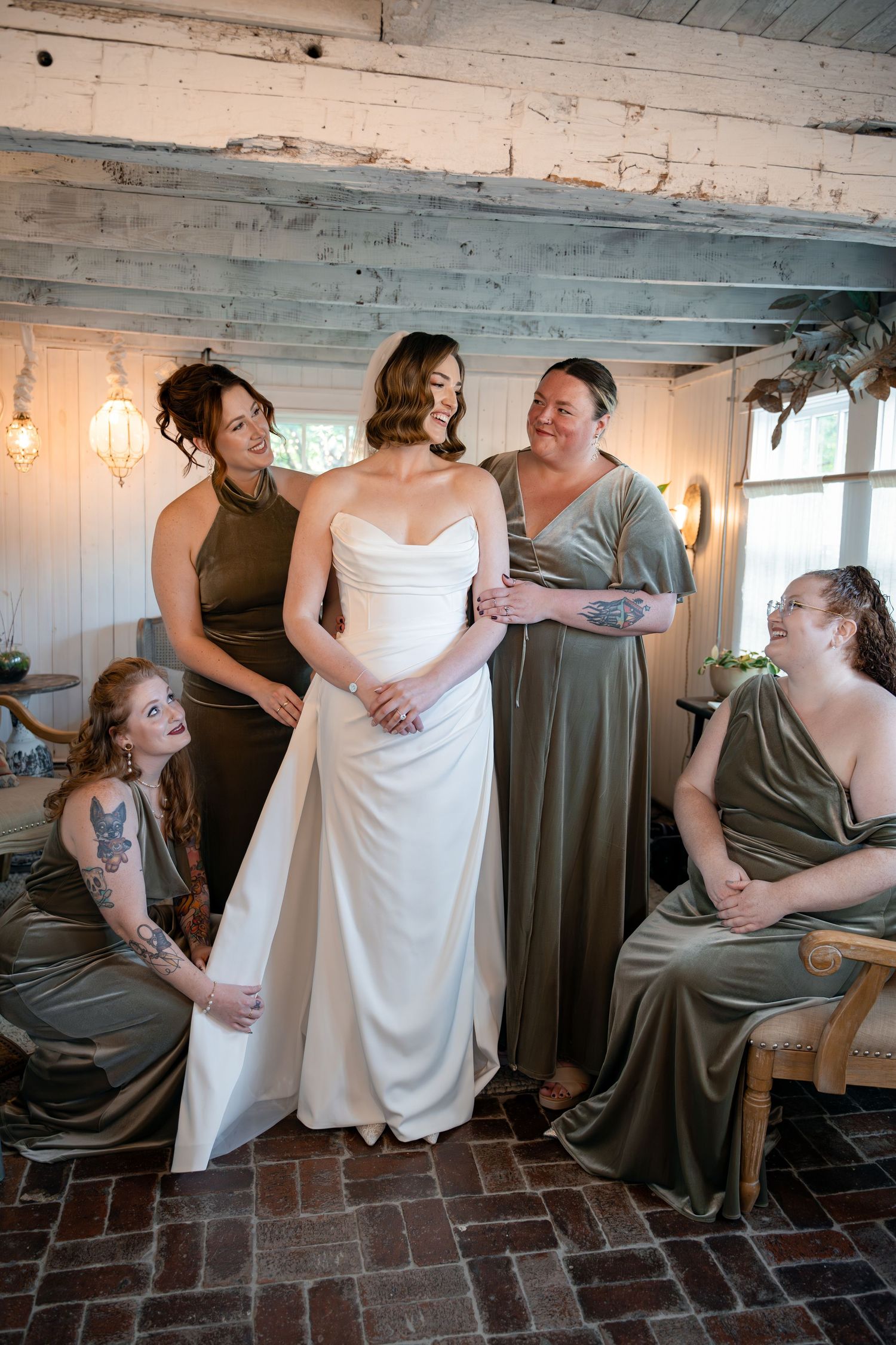 Bride in white gown with bridesmaids in olive green dresses laughing together in rustic venue at Terrain at Styers, PA