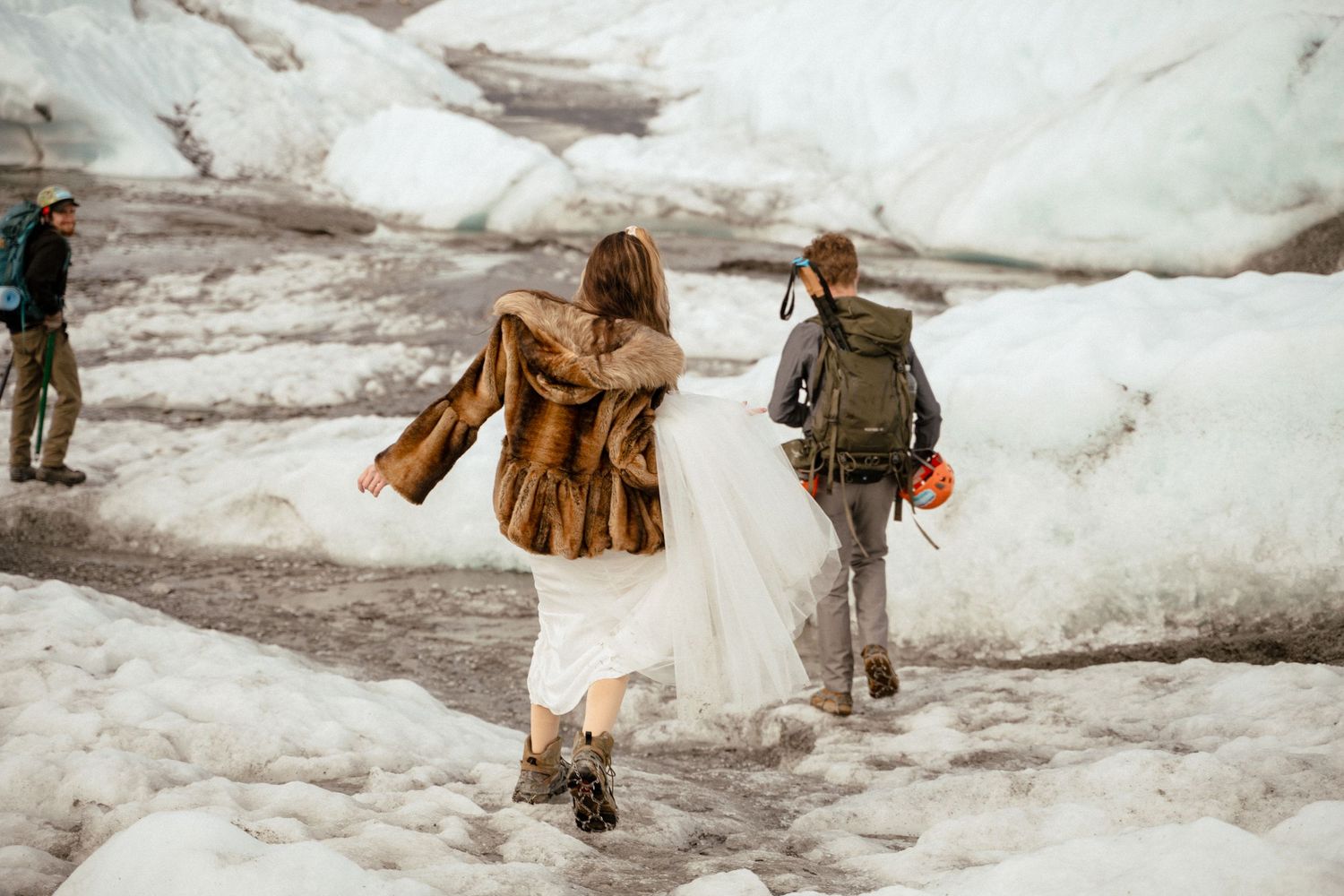 Bride in fur coat and groom with backpack walking on glacier during winter elopement wedding adventure.