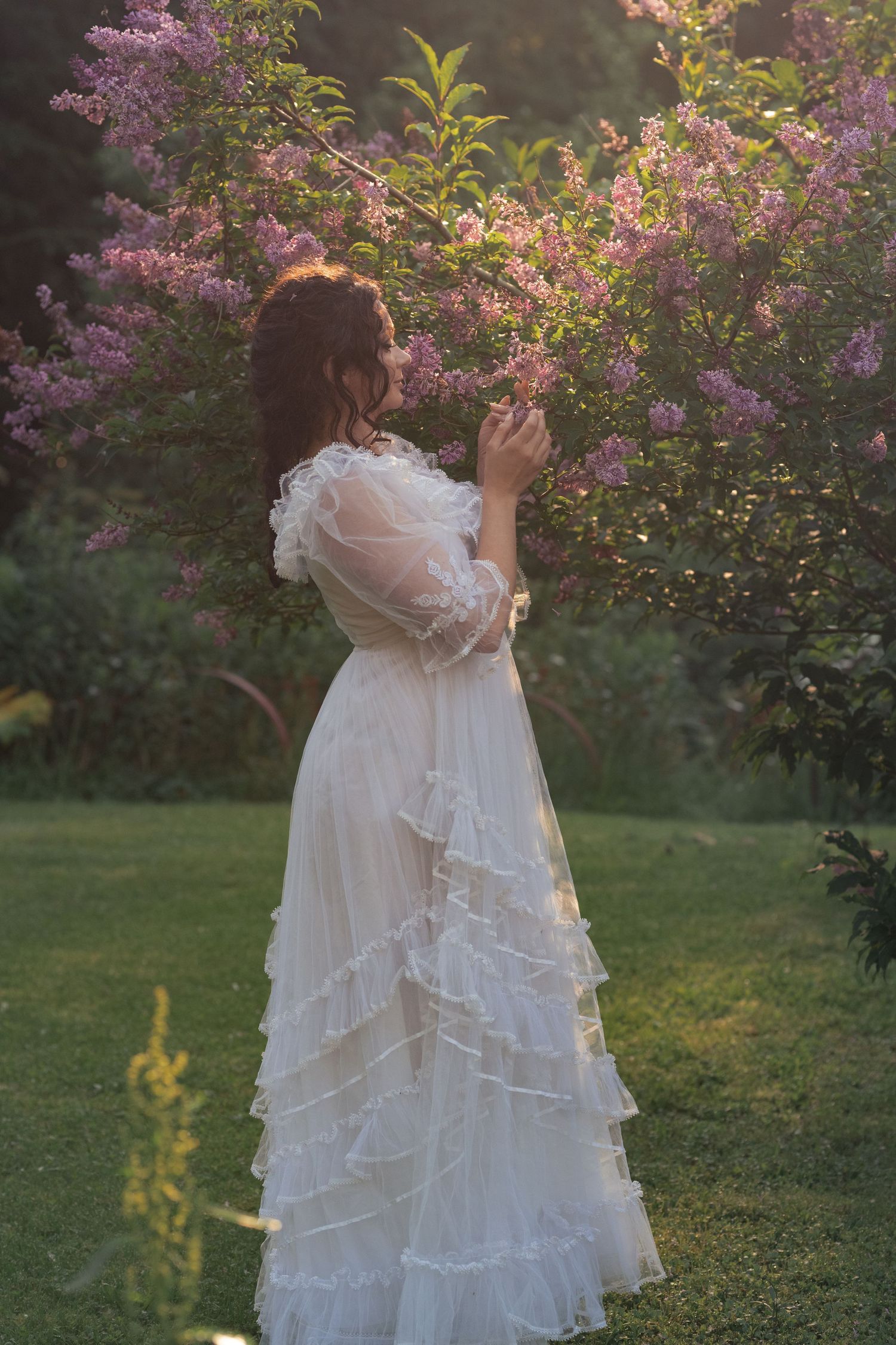 Woman in white ruffled Victorian dress standing beside blooming lilac bushes in a sunlit garden.