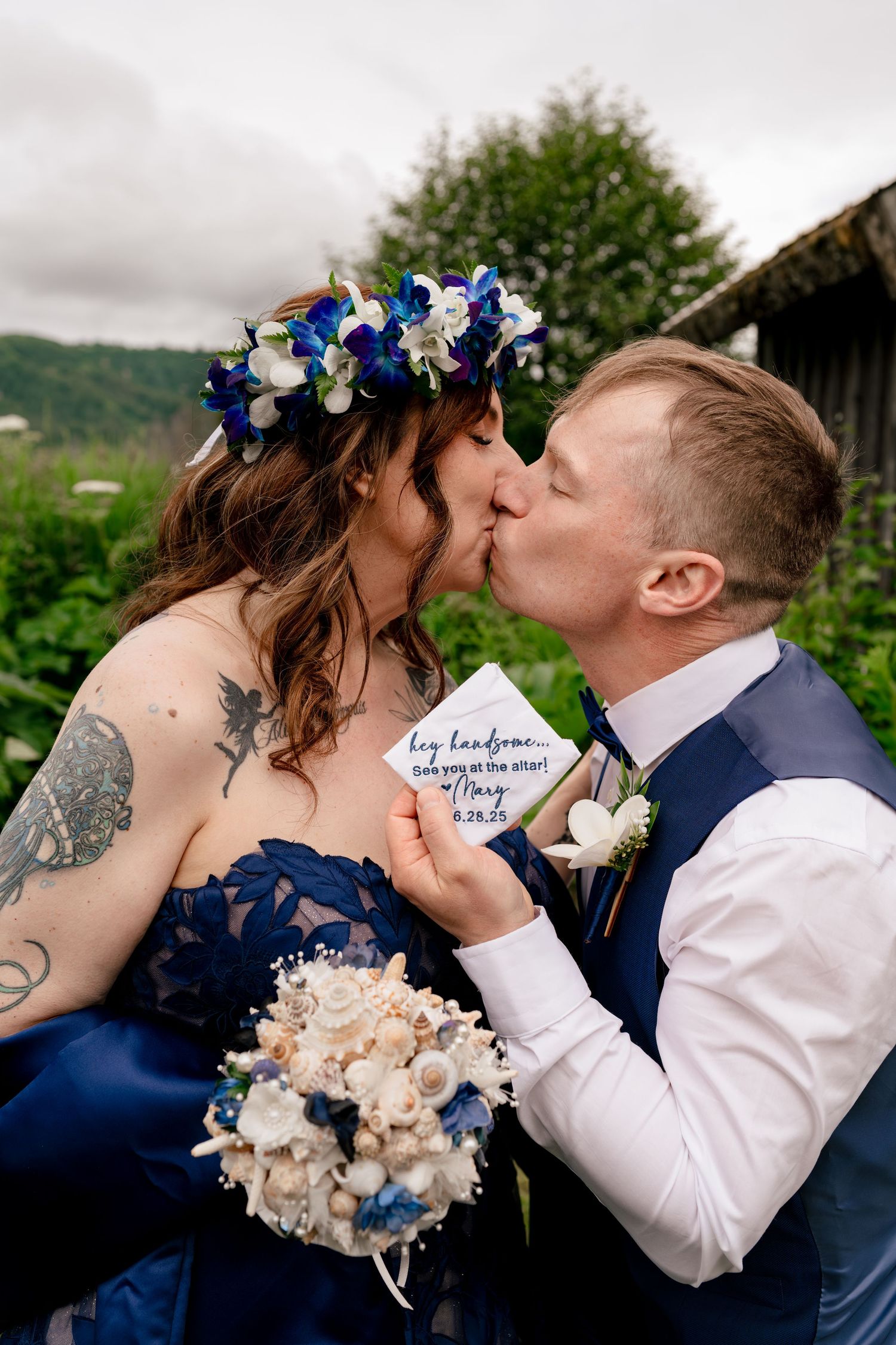 Bride and groom share a kiss outdoors, bride wearing blue floral crown holding bouquet, groom in navy vest.