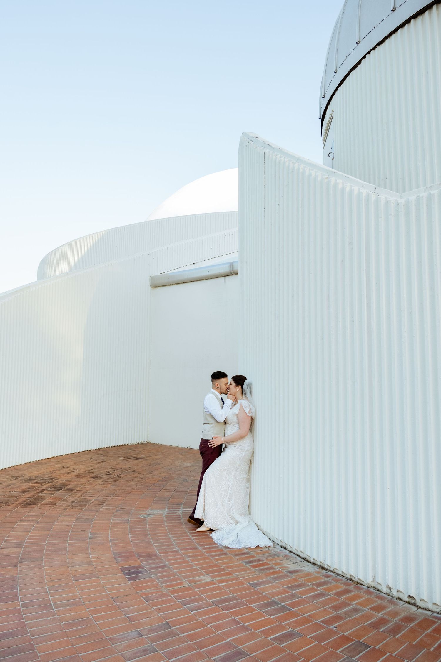 Bride and groom share a romantic moment against a curved white architectural wall on a brick courtyard.