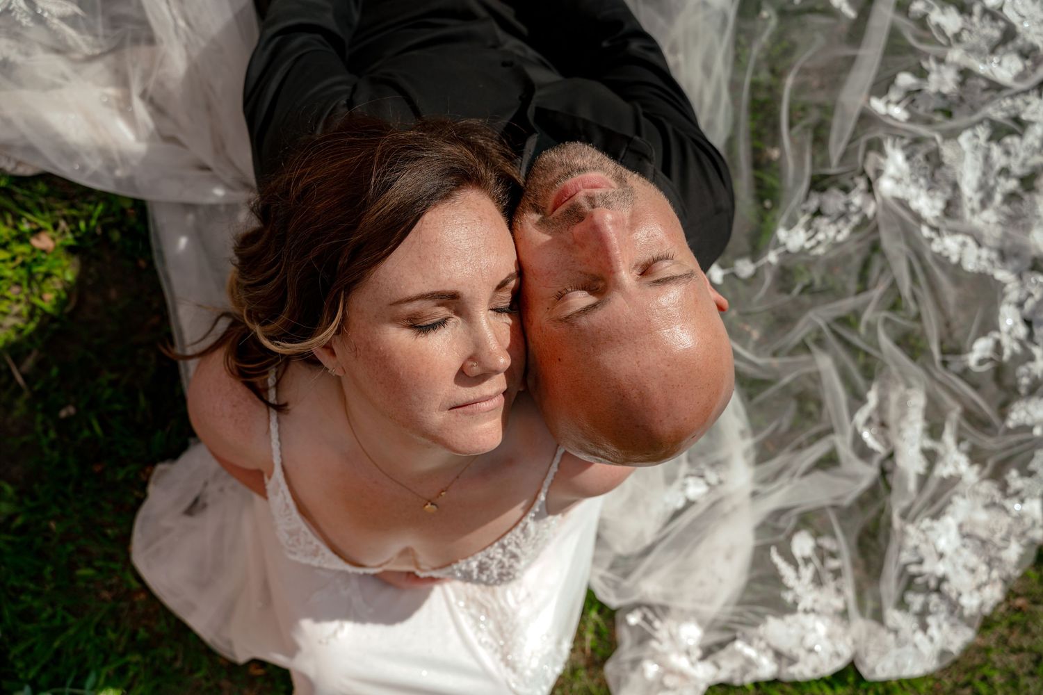 Aerial view of a bride and groom lying on grass, eyes closed, in wedding attire, sharing a peaceful moment.