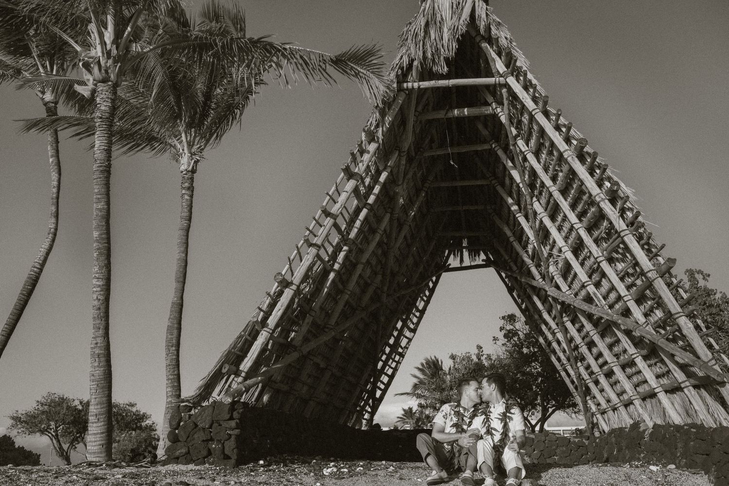 Black and white photo of a traditional A-frame wooden structure with palm trees and people in traditional attire nearby.