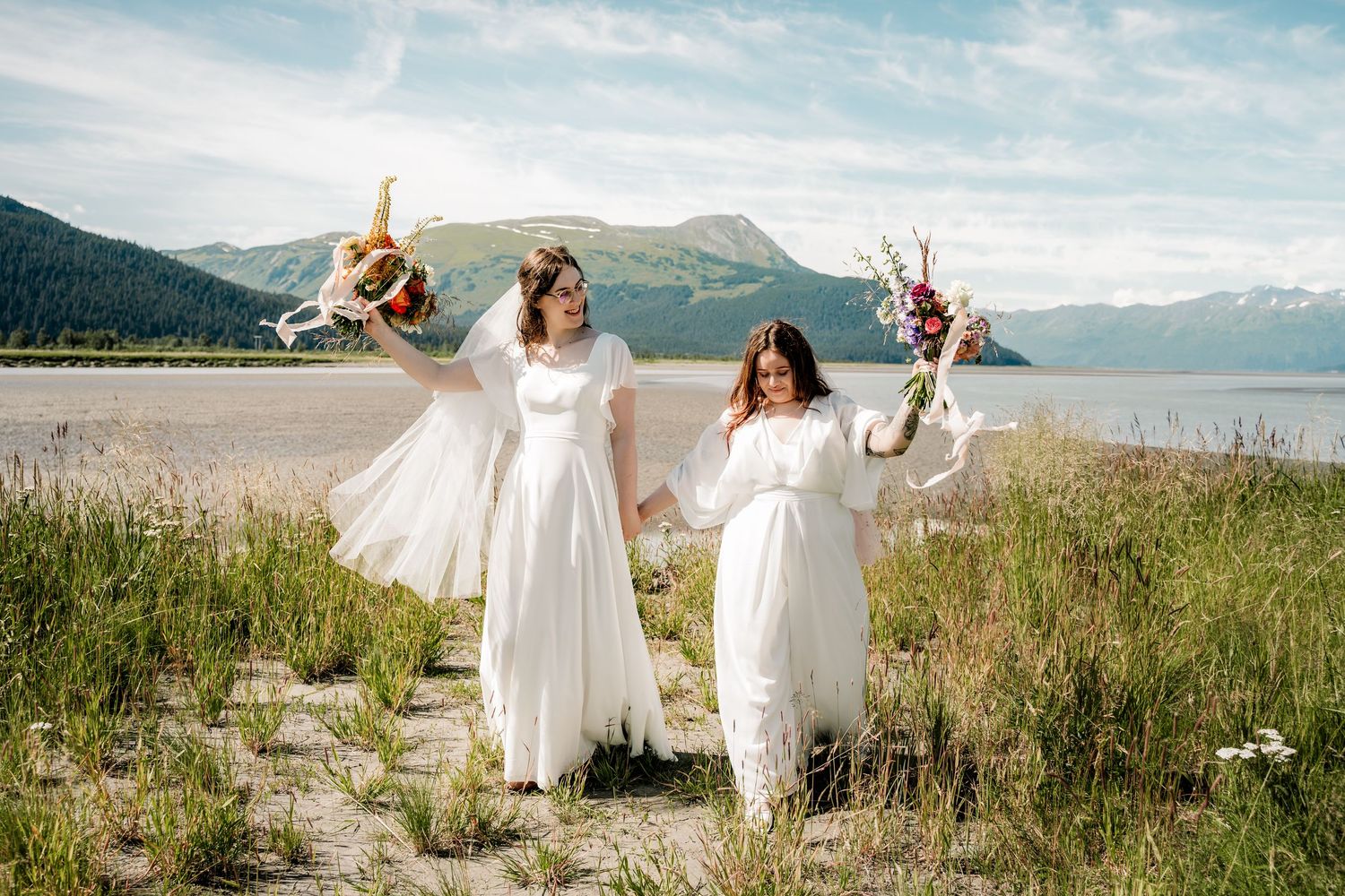 People in flowing white dresses dance and twirl together in a scenic mountain meadow with lake views in the background.