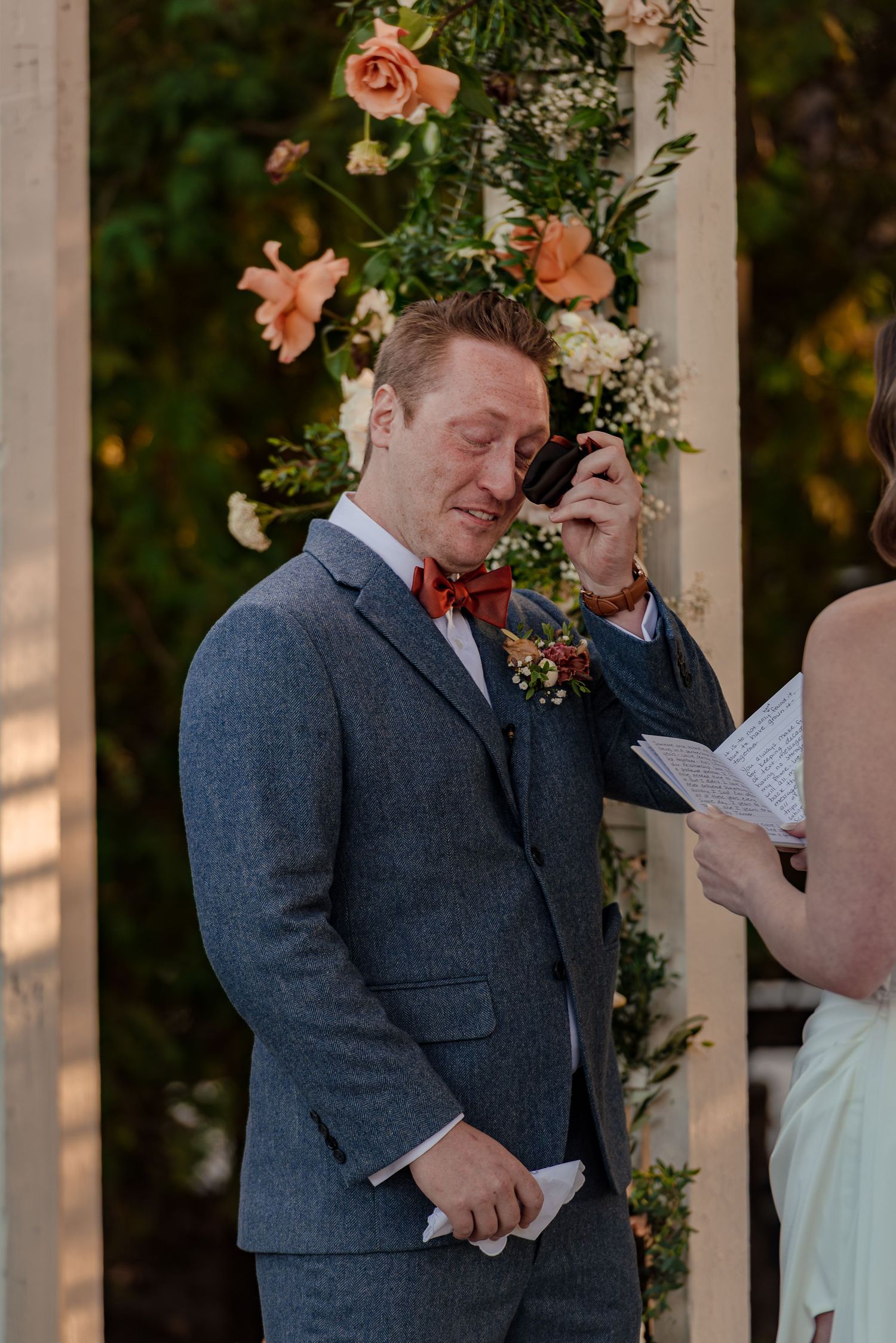 Emotional groom wiping tears during outdoor wedding ceremony, wearing blue suit and rust bow tie with floral arch backdrop.