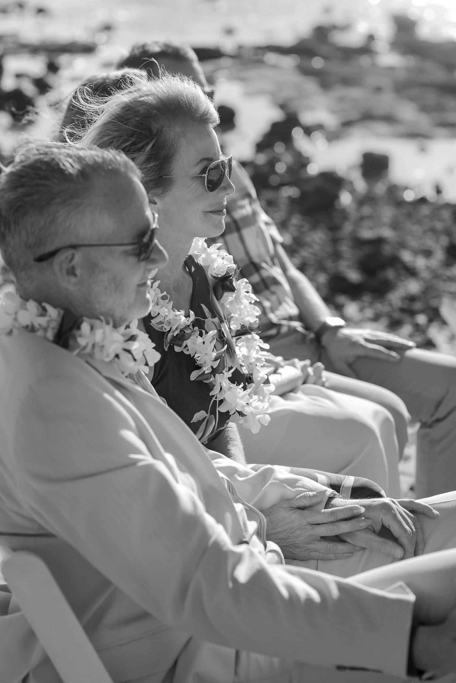 Elderly couple wearing flower leis seated outdoors at a Hawaiian beach ceremony in black and white photo.