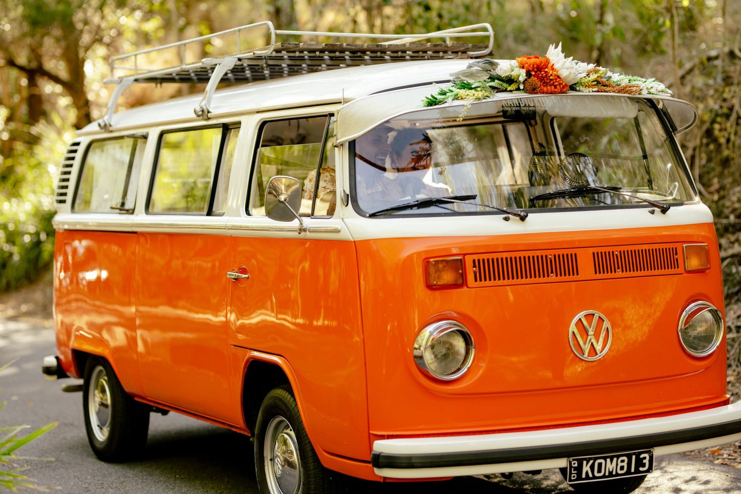 Vintage orange VW Kombi van decorated with flowers on roof, parked on a tree-lined road.