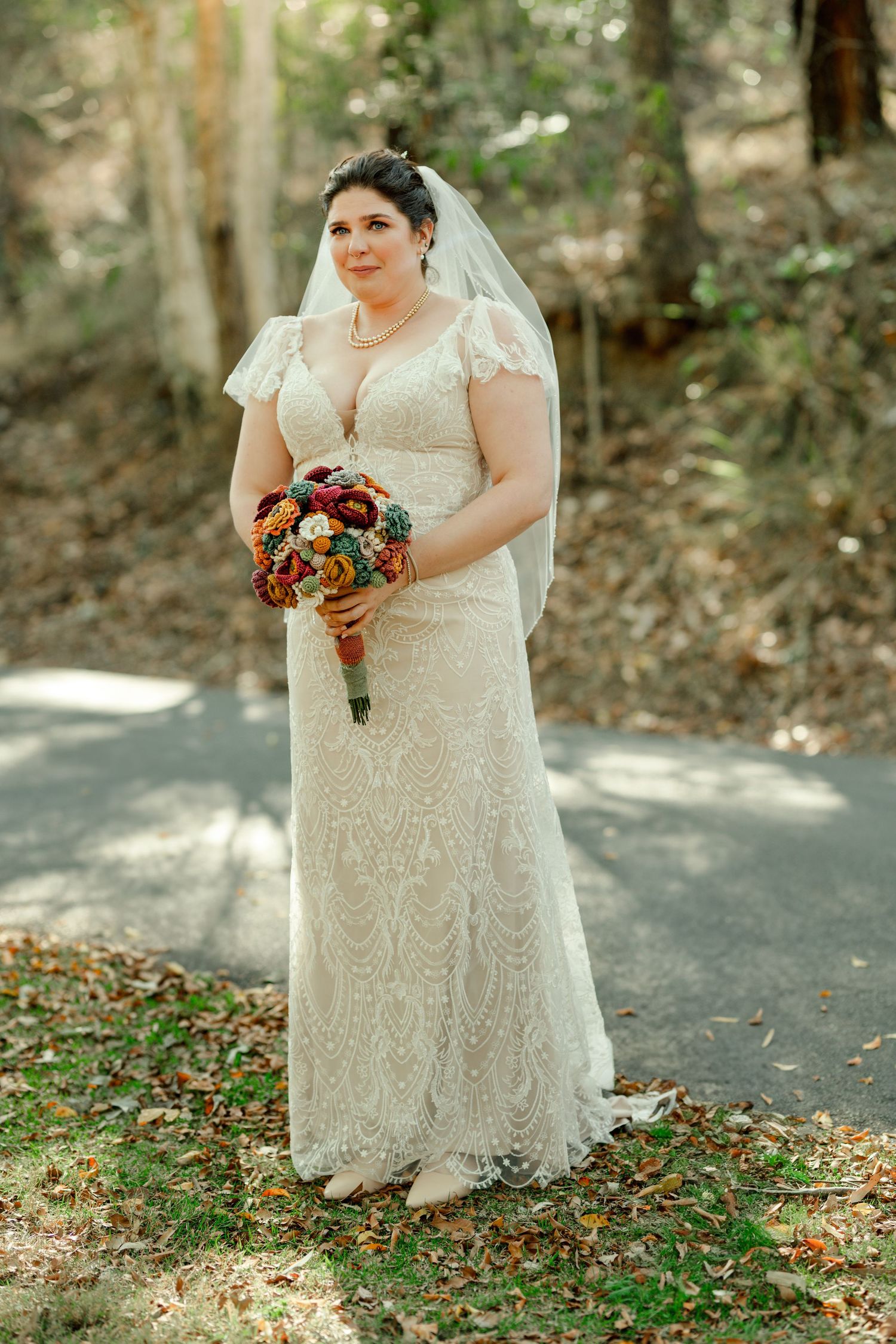 Bride in lace wedding gown and veil holds colorful bouquet outdoors in wooded setting.