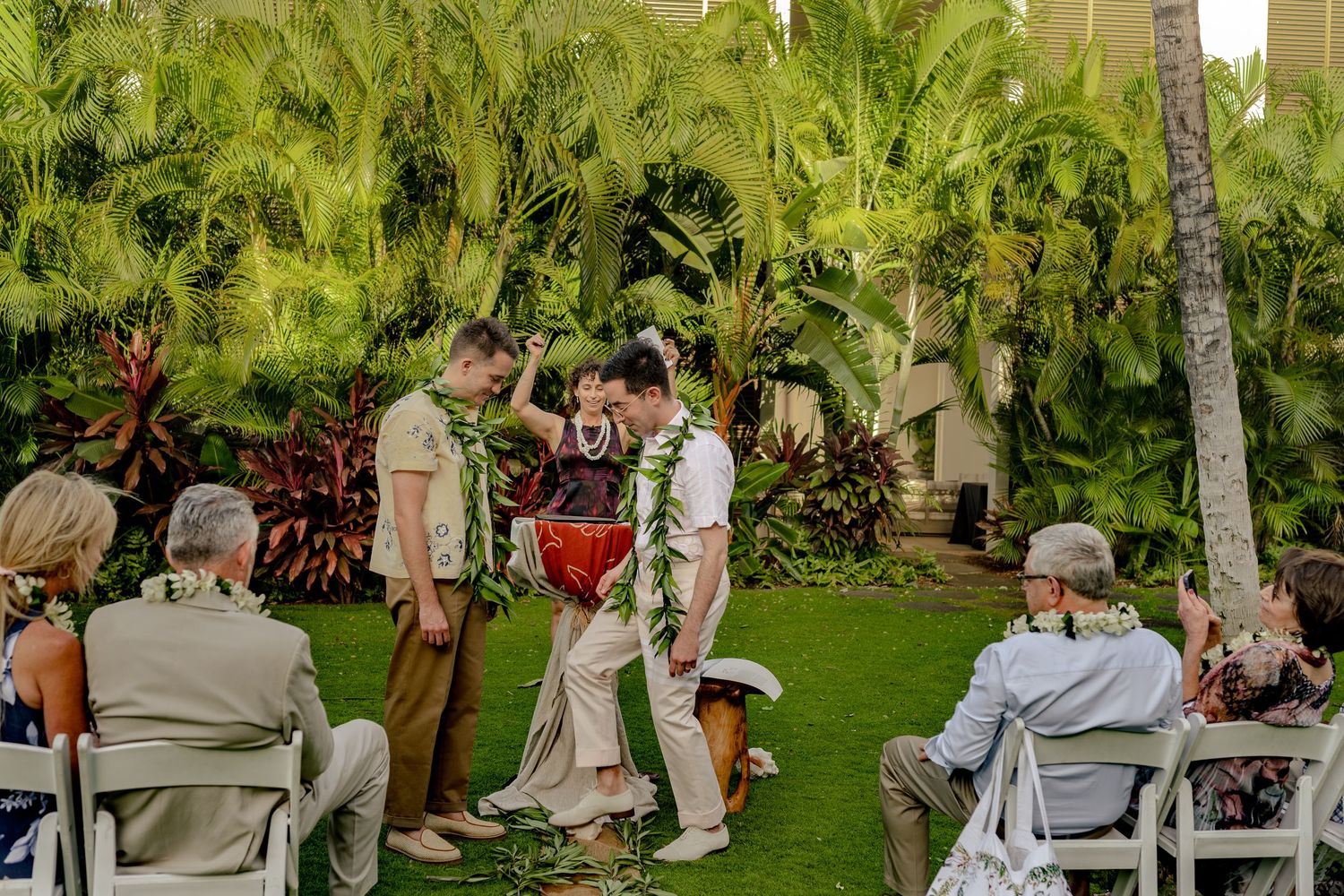 Tropical outdoor wedding ceremony with couple exchanging leis amid lush palm trees and seated guests on white chairs.