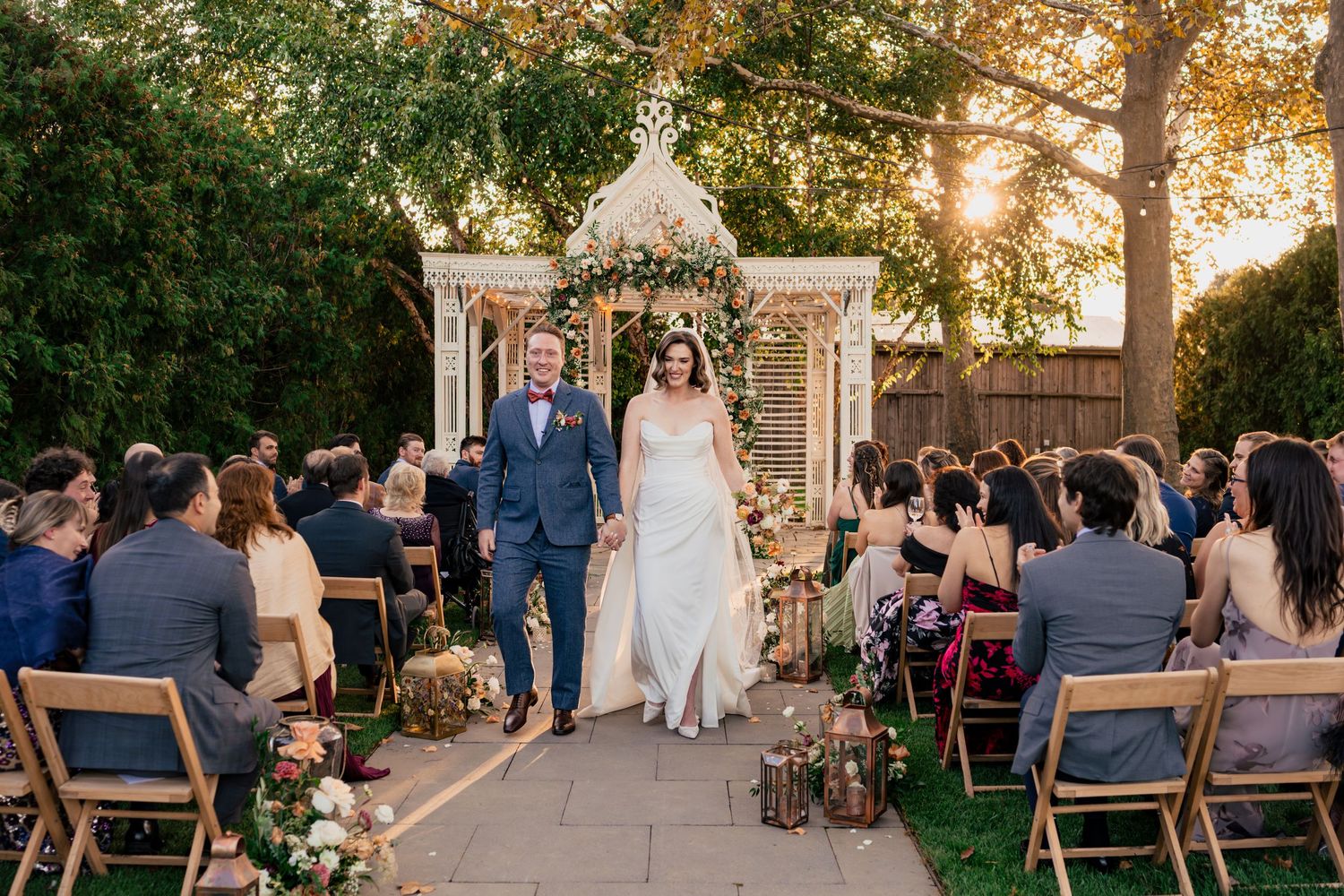 Bride and groom walk down outdoor aisle at sunset wedding ceremony with guests seated on wooden chairs.