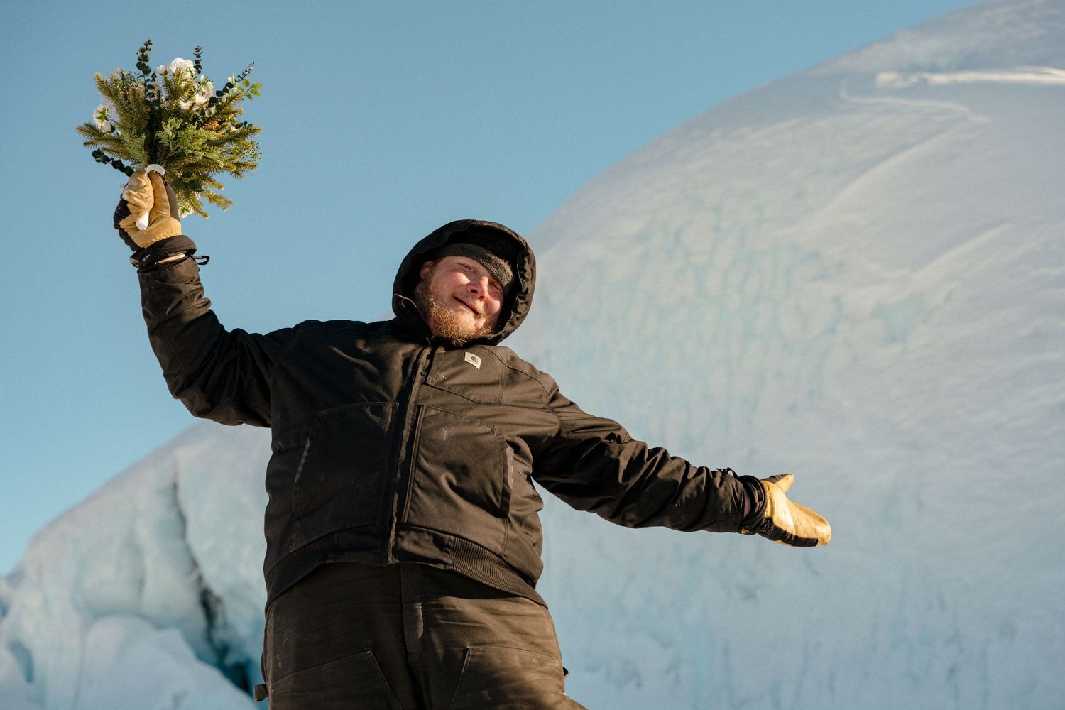 Person in black jacket holding wildflowers with arms outstretched, celebrating among icy glaciers in a snowy landscape.