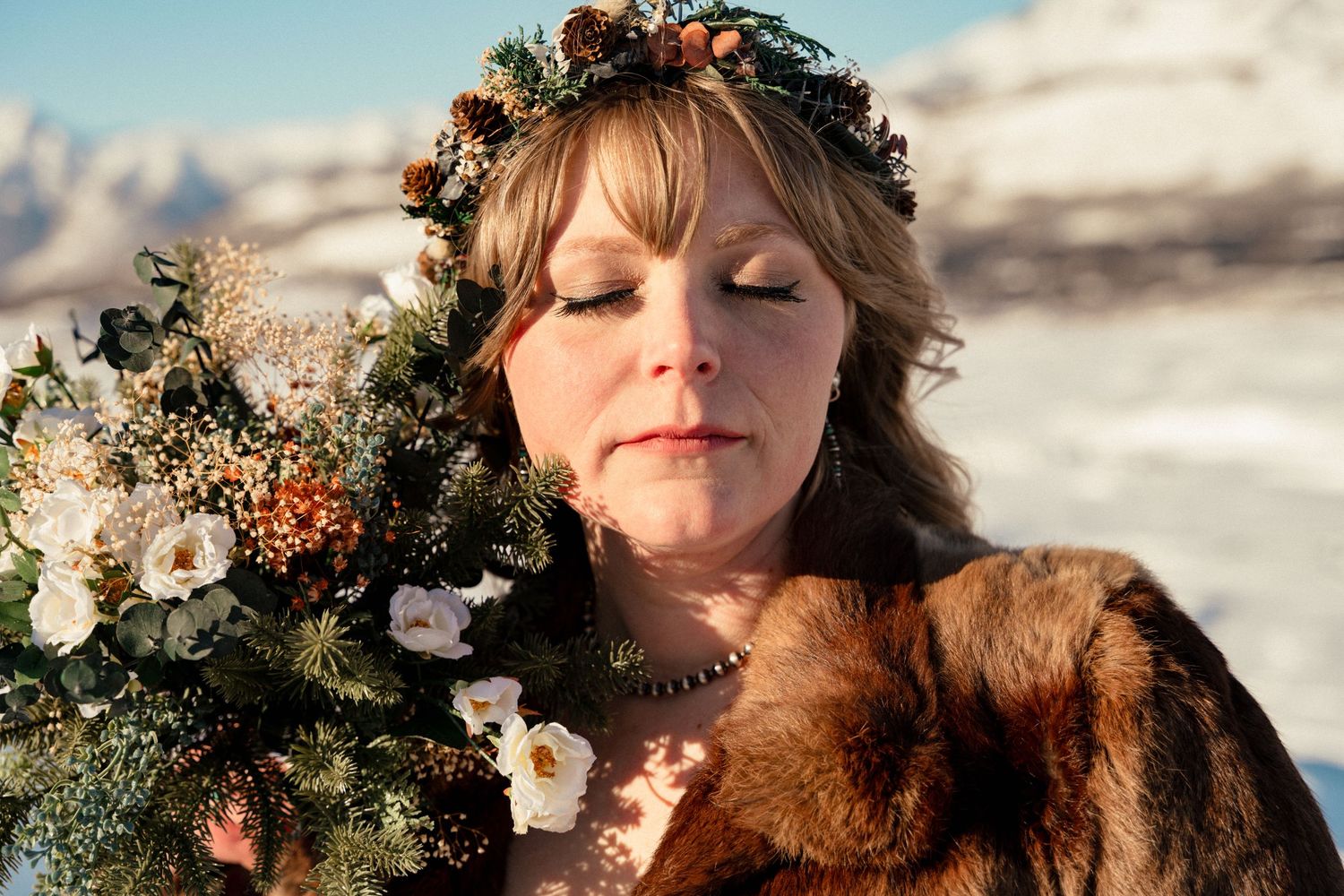 Woman in fur coat and floral crown holds winter bouquet with snowy mountains in background.