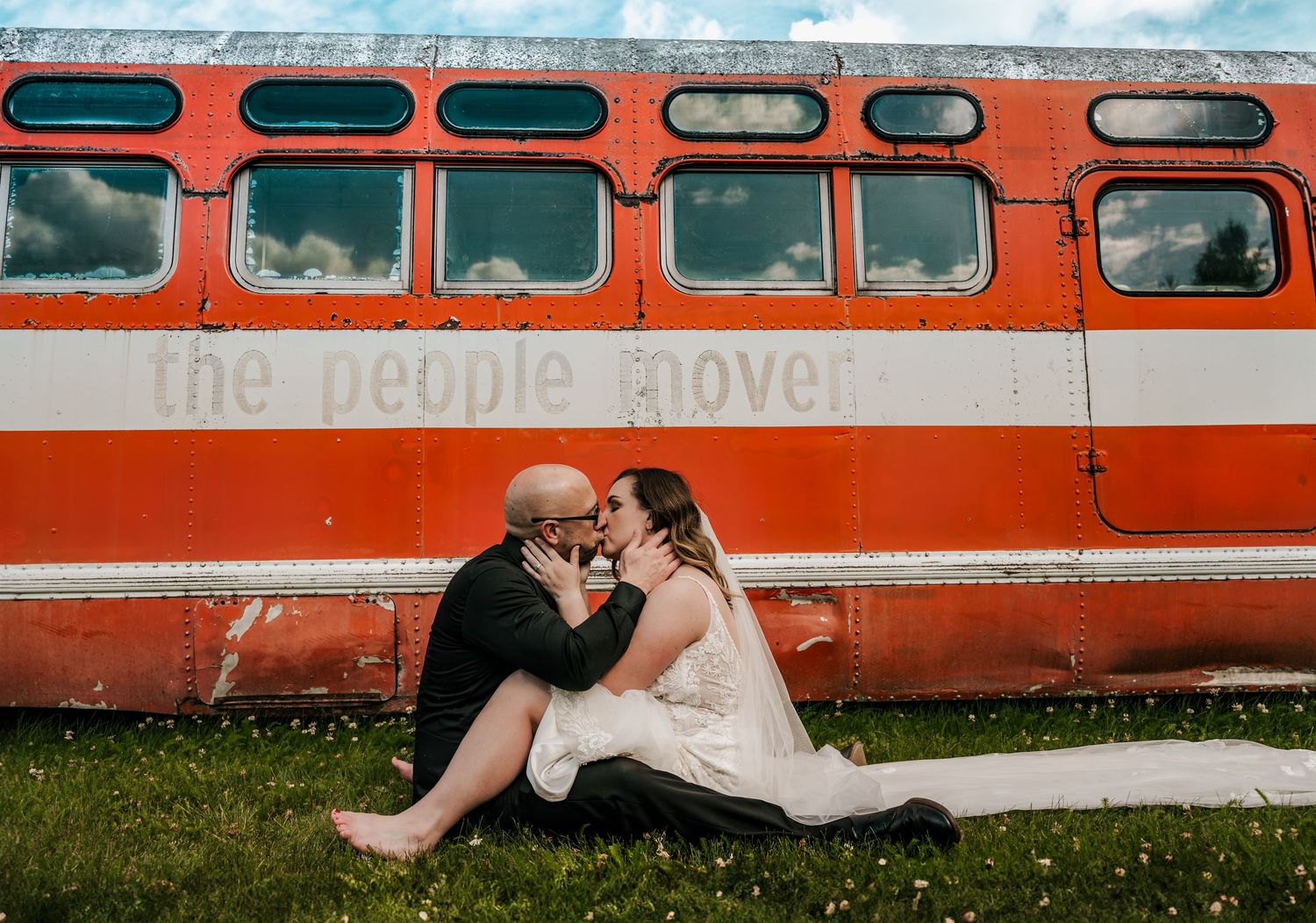 Couple sharing a romantic kiss in wedding attire seated in front of a vintage red People Mover bus in Wasilla Alaska