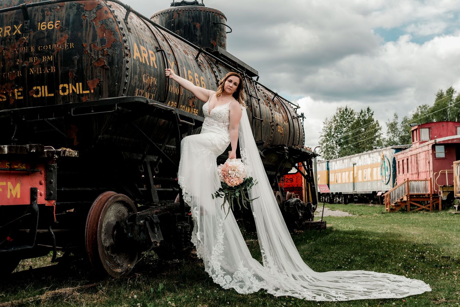 Bride in a flowing white lace gown poses elegantly beside a vintage train at an outdoor railway museum.
