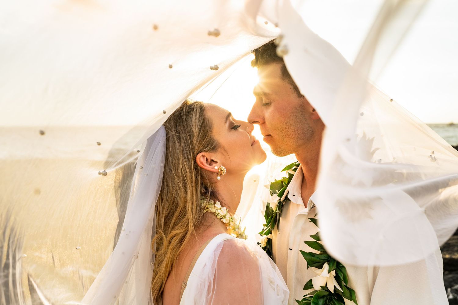Intimate wedding moment captured under flowing veil with sunlight streaming through.