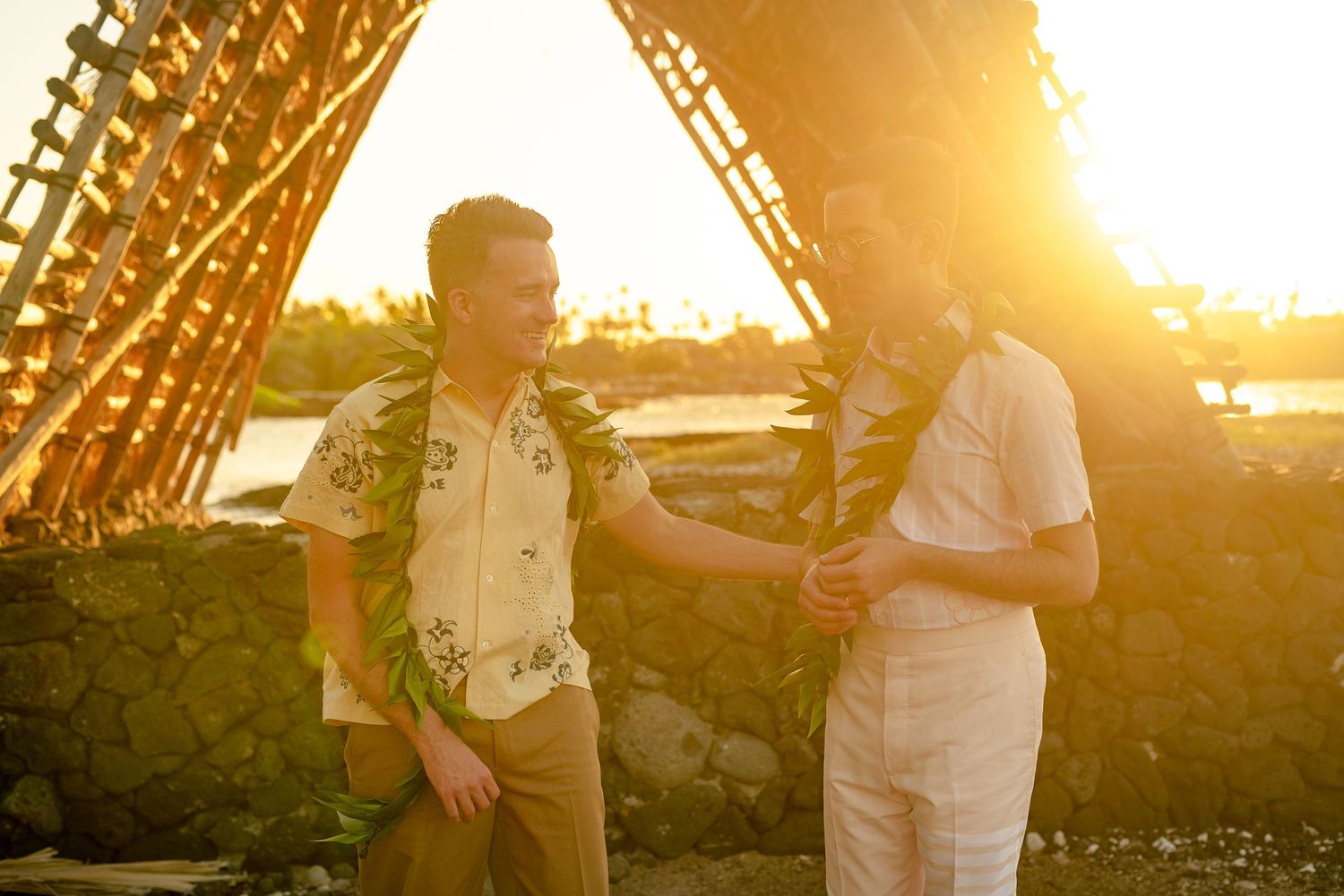Two grooms exchanging smiles outdoors near a bamboo structure bathed in warm golden sunset light.