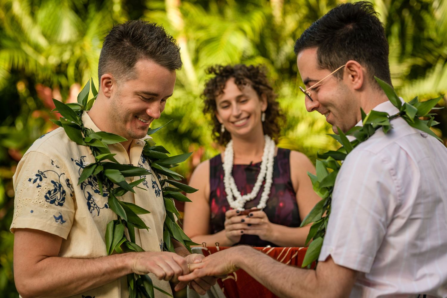 Two grooms exchange rings at a Hawaiian wedding ceremony, wearing green leaf leis, with a smiling officiant behind them.