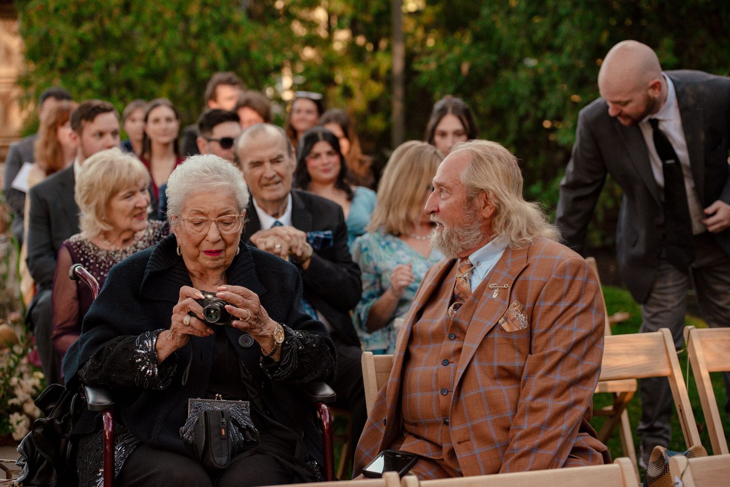 Elderly guests seated at an outdoor wedding ceremony, woman photographing with camera, man in brown tweed suit.