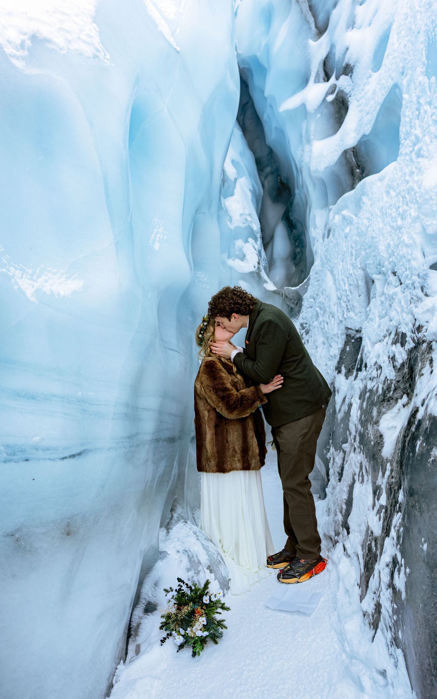 Couple sharing a romantic kiss inside a stunning ice cave, bride in white dress and fur coat with a floral bouquet on snow.