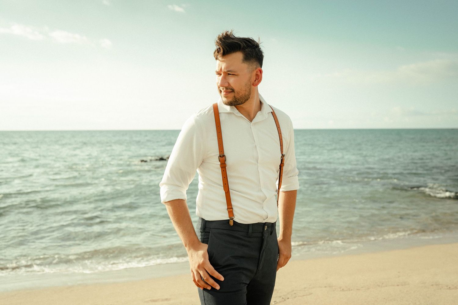 Man in white shirt and brown suspenders standing on a sandy beach with ocean horizon in background.