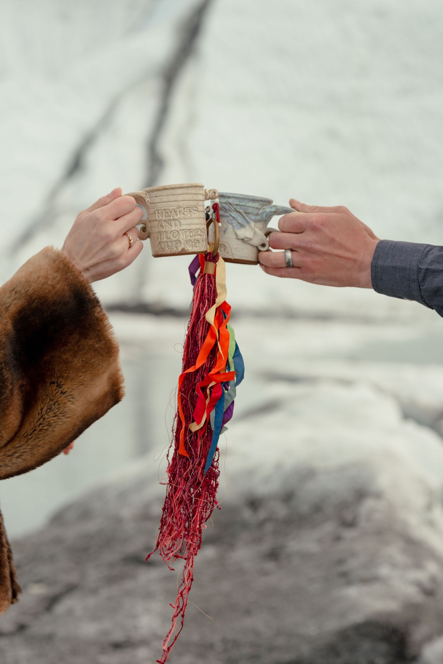 Two hands clinking rustic ceramic mugs with colorful fringe ribbons hanging between them against a snowy backdrop.
