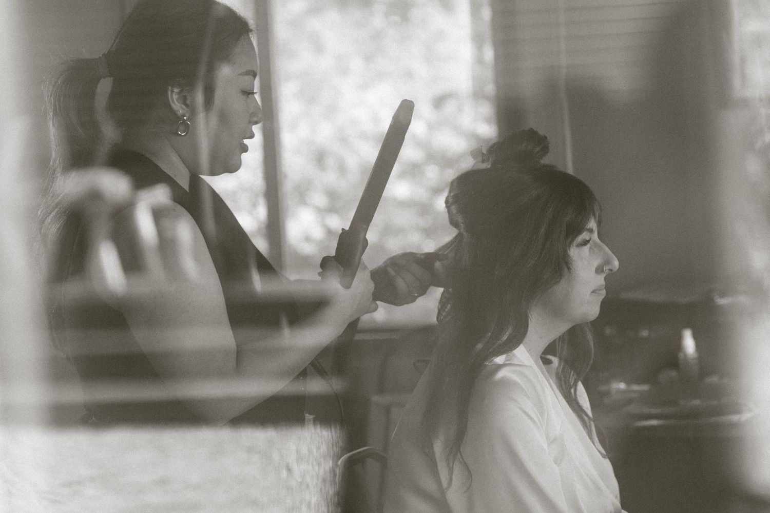 A hairstylist uses a flat iron on a seated woman's hair in a softly lit room, captured in black and white.