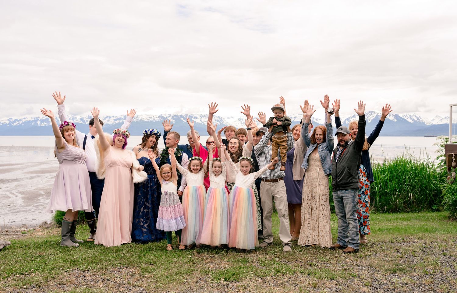 Wedding party celebrates outdoors near Alaska beach with mountains, all raising hands joyfully in colorful formal attire.