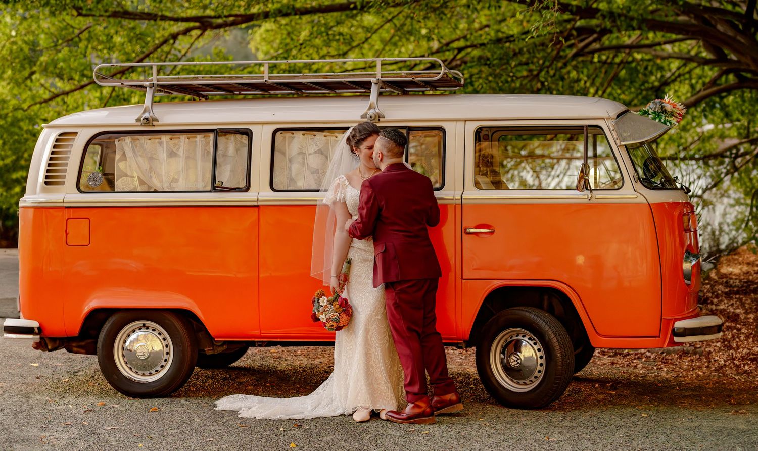 Bride and groom kissing in front of a vintage orange VW camper van at their wedding.