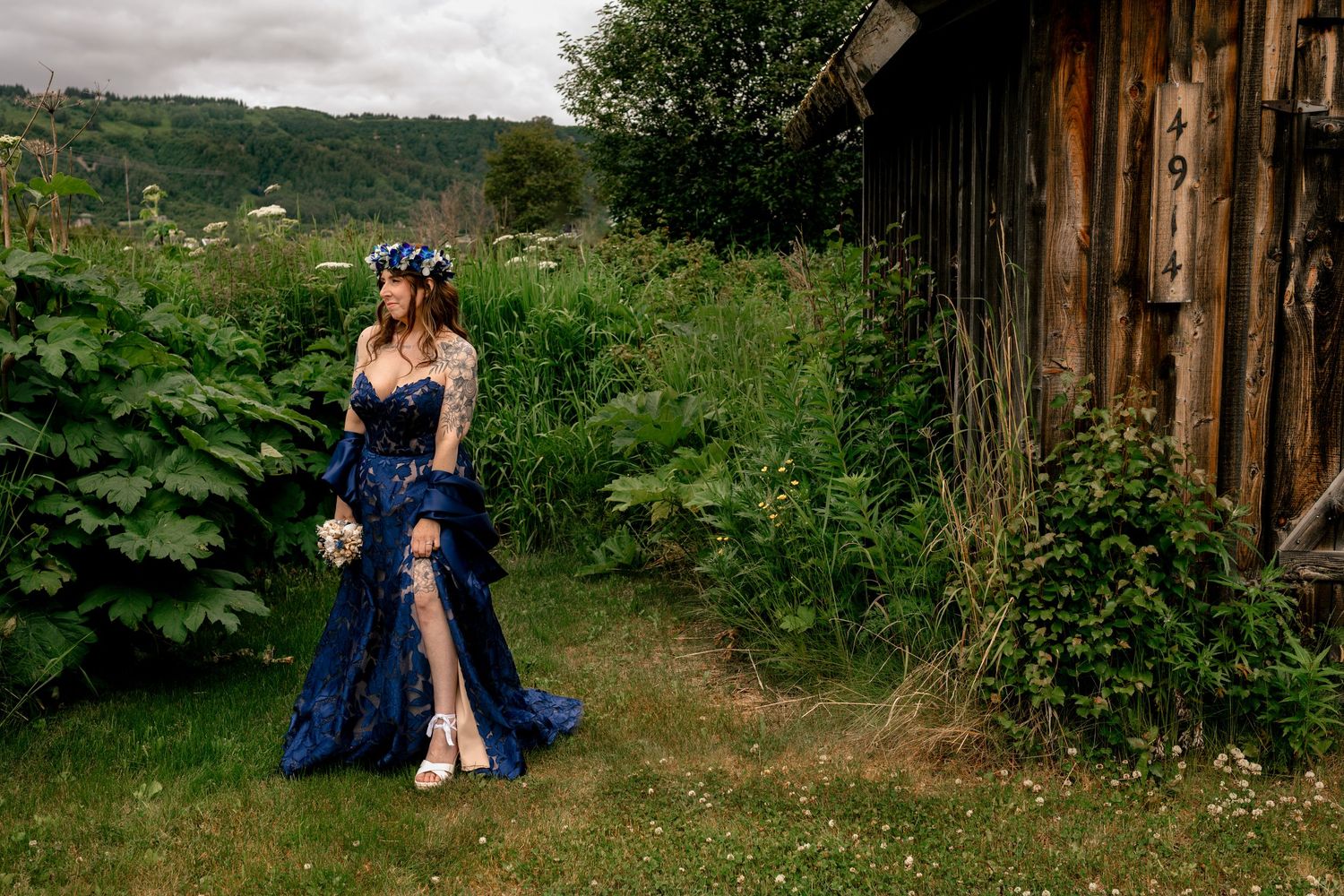 Woman in navy blue gown with floral crown poses near rustic barn surrounded by lush greenery.
