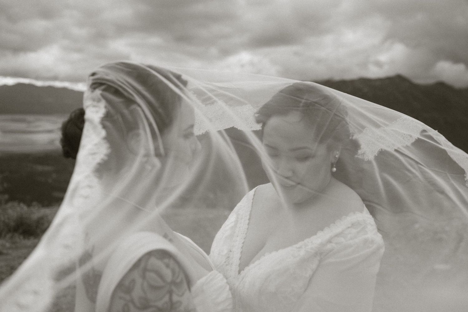 Black and white photo of two brides under a flowing veil with mountains in the background.