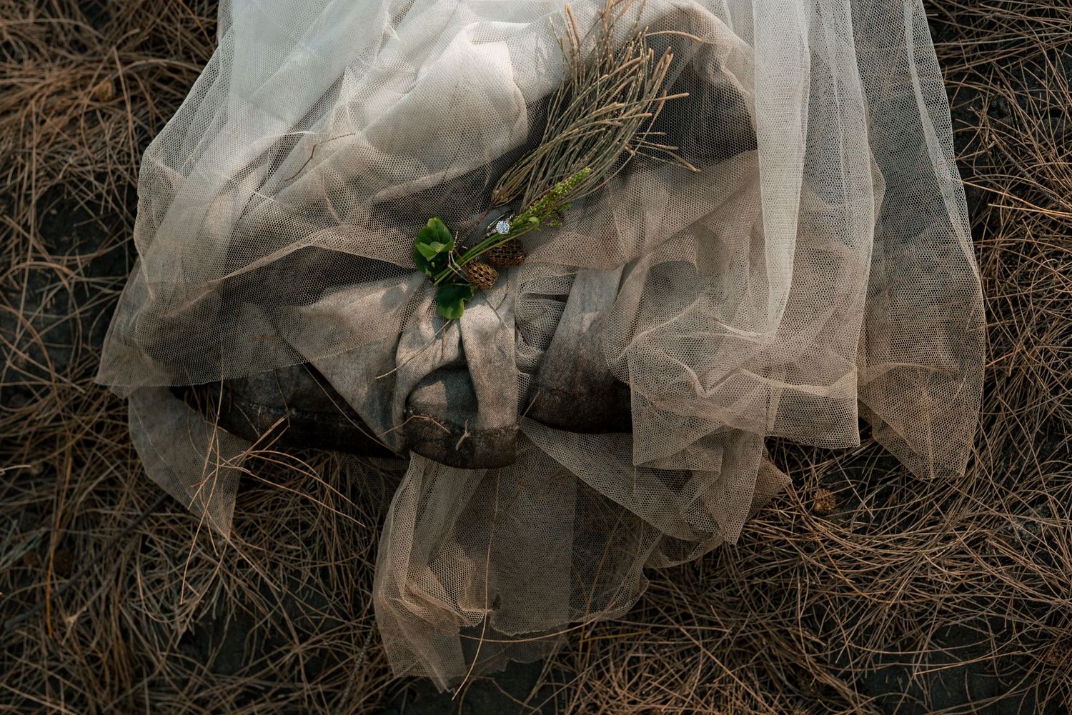 Wedding dress and veil details laying on rustic ground with greenery accents.