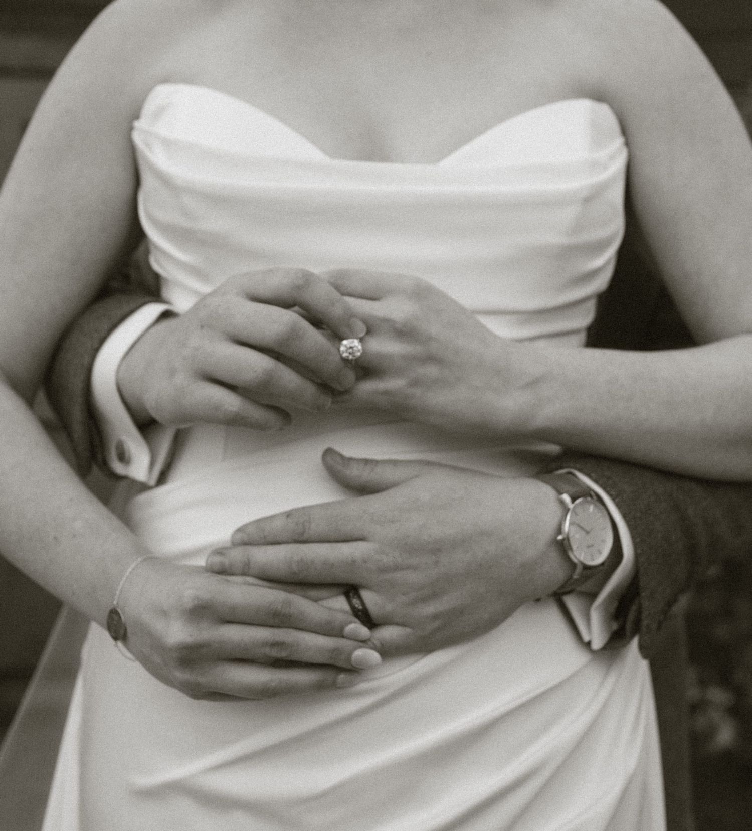Black and white photo of bride and groom embracing, showing wedding rings and watch on clasped hands.