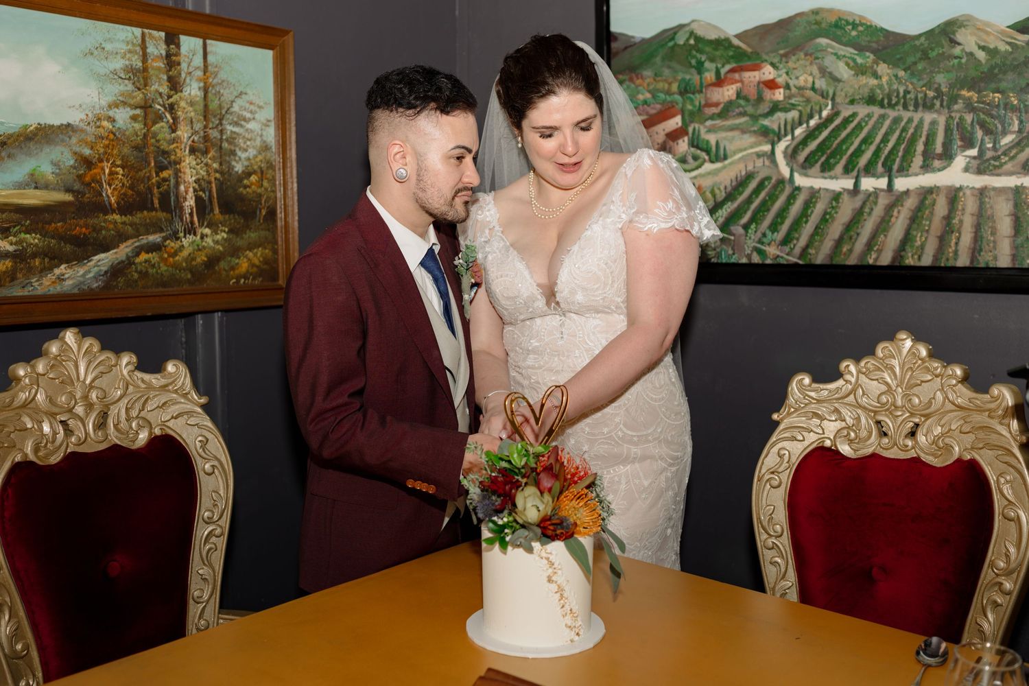 Bride and groom cutting white wedding cake at decorated table with ornate chairs and vineyard paintings on wall.