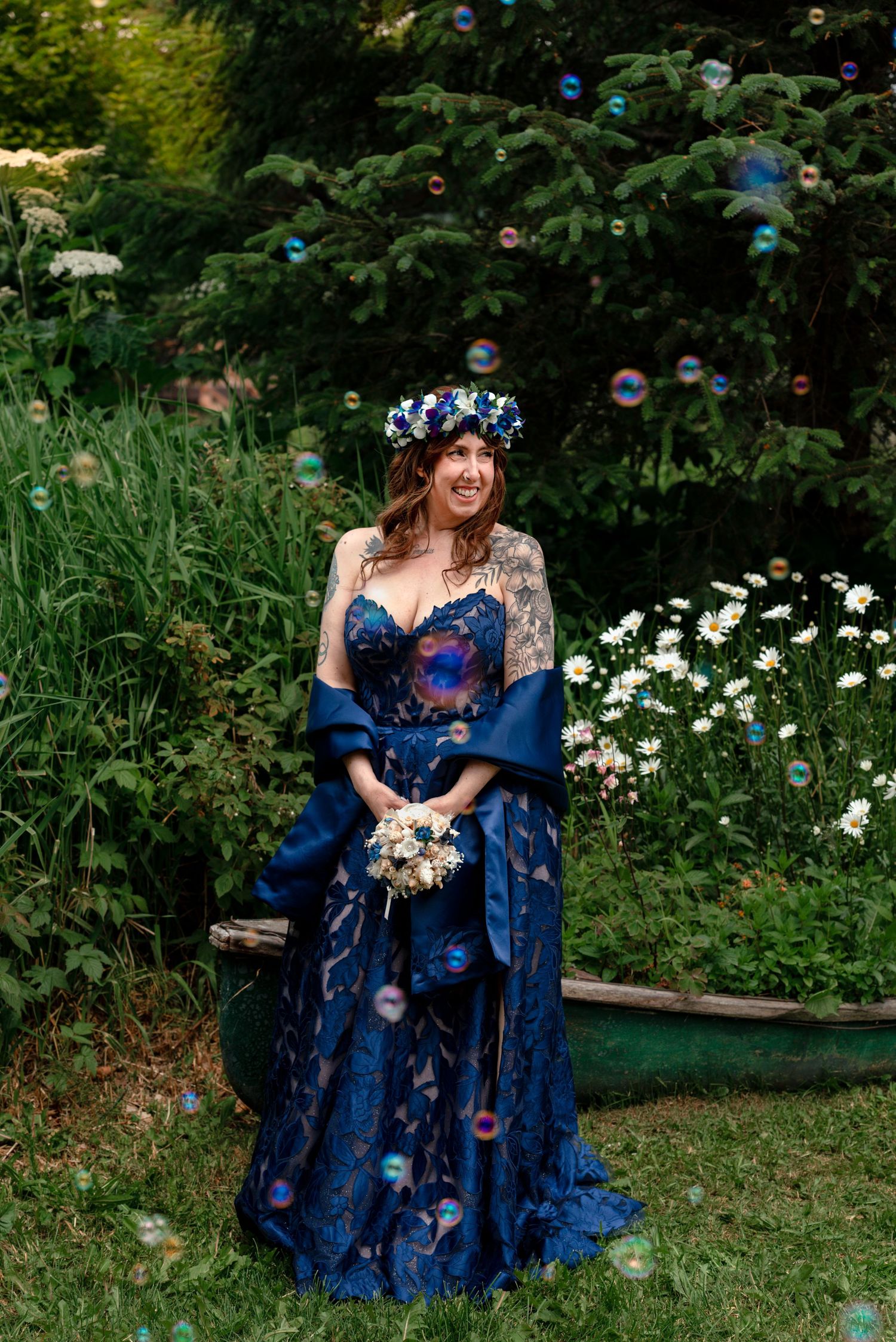 Smiling bride in blue floral gown and flower crown holds bouquet surrounded by bubbles in lush garden.