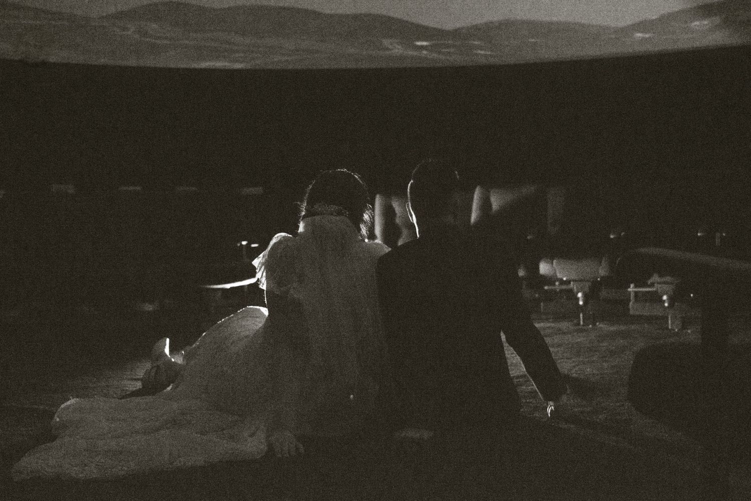 Black and white photo of a couple sitting together outdoors at night with mountains in the background.