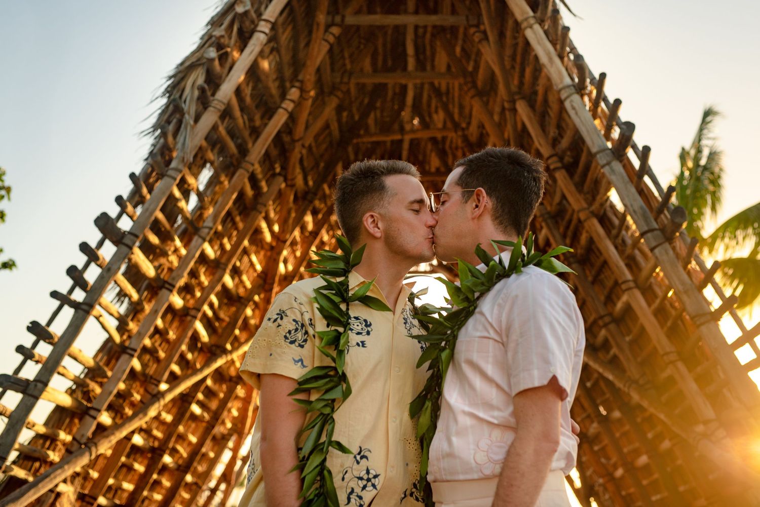 Two grooms share a kiss wearing floral leis under a bamboo structure at golden hour in Hawaii.