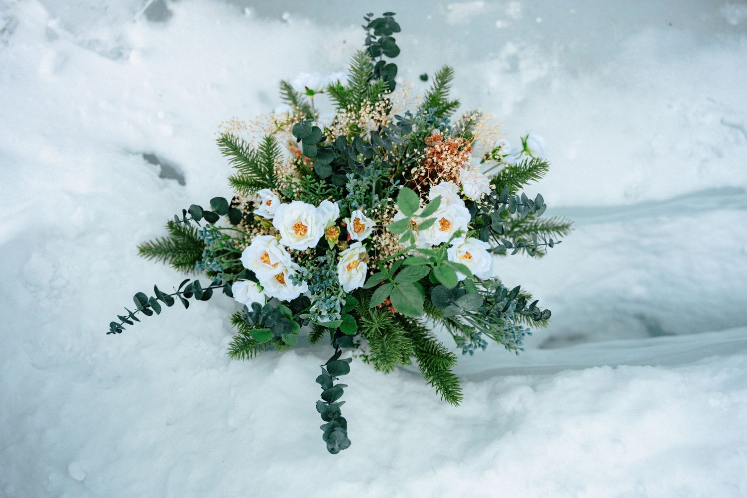 Winter wedding bouquet with white flowers and greenery resting on snow.