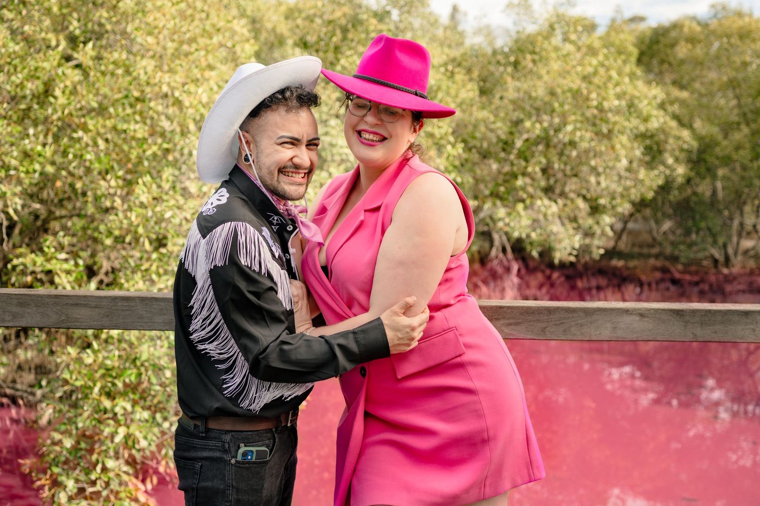Two women in pink and black cowboy hats dance and smile outdoors near lush greenery and a pink smoky background.