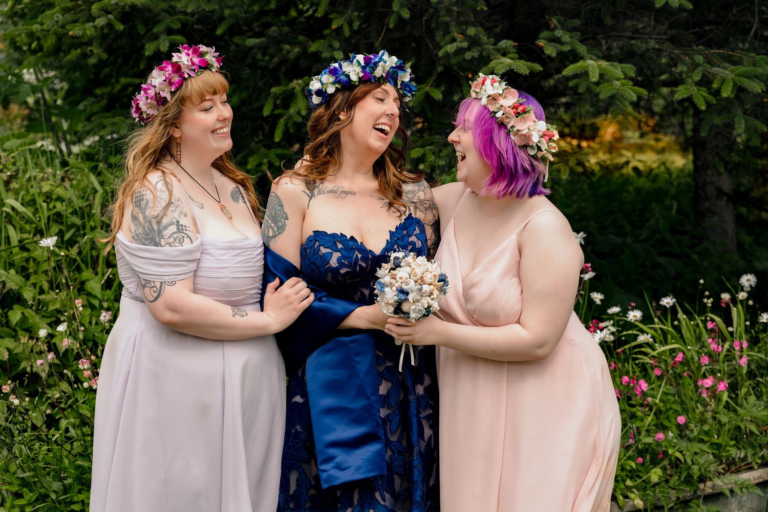 Three women in floral crowns laugh together outdoors, one in blue wedding dress holding bouquet, two in bridesmaid gowns.
